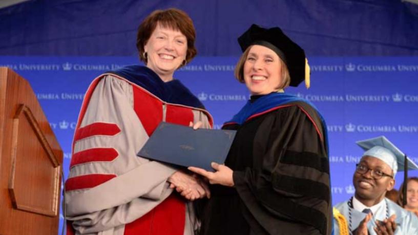 One woman hands another woman an award while a male student, in the background, applauds