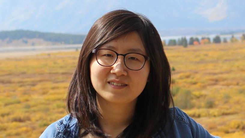 Photo of Dr. Tian. She is standing outside under a blue sky. She is smiling and wearing blue and yellow clothing.