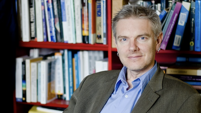 Photo of Prof. Simon Billinge. He is sitting in front of a bookcase. He is wearing a blue shift and a brown jacket.