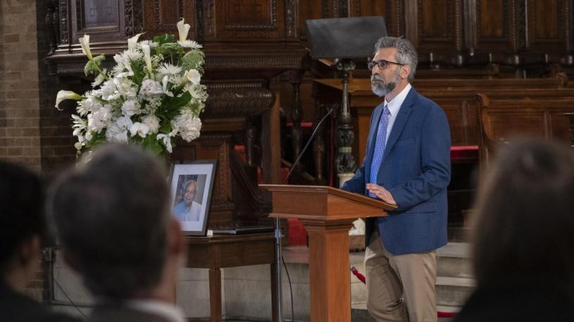 A photo of Marc Spiegelman. He is wearing a blue suit and tie and is standing at a podium in the front of a chapel