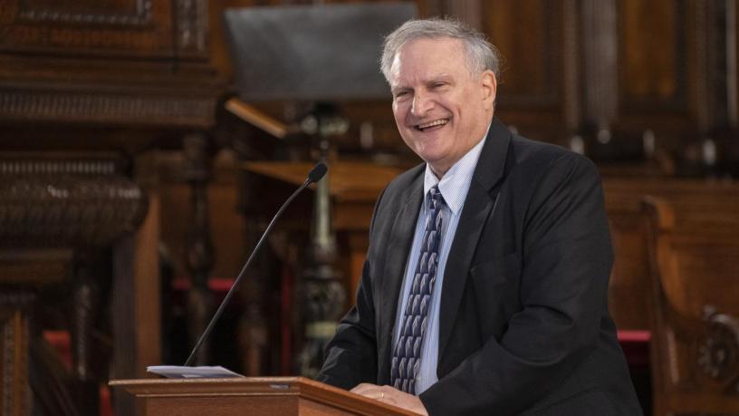 A photo of Prof. Gerlad Navratil. He is wearing a black suit and tie and is standing at a podium in the front of a chapel.