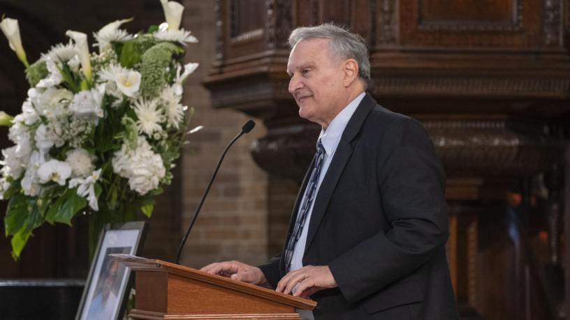 A photo of Prof. Gerlad Navratil. He is wearing a black suit and tie and is standing at a podium in the front of a chapel.