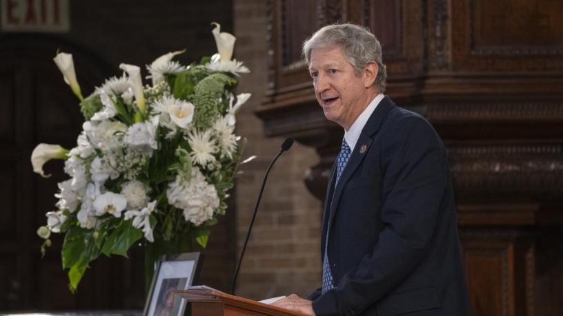 A photo of Prof. David Keyes. He is wearing a black suit and tie and is standing at a podium in the front of a chapel.