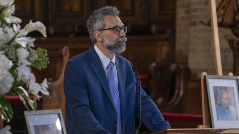 A photo of Marc Spiegelman. He is wearing a blue suit and tie and is standing at a podium in the front of a chapel