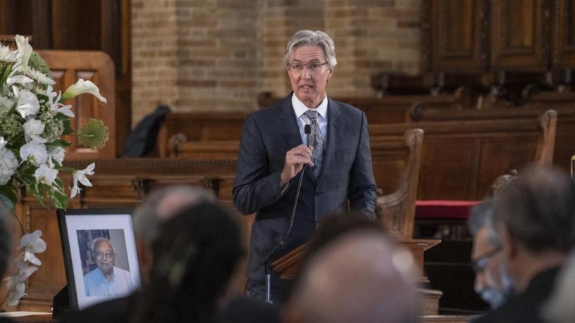 A photo of Dr. Ralph Izzo. He is wearing a grey suit and tie and is standing at a podium in the front of a chapel.