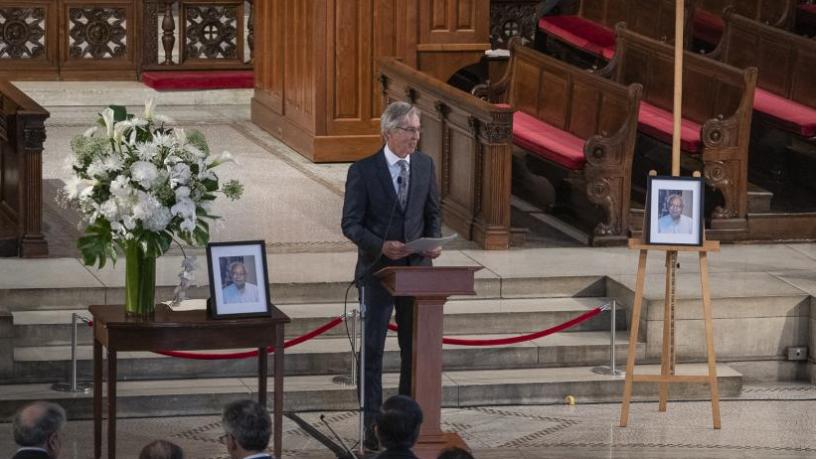 A photo of Dr. Ralph Izzo. He is wearing a grey suit and tie and is standing at a podium in the front of a chapel. This photo is taken at a great distance and you can see the largeness of the chapel,