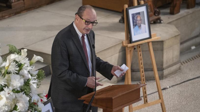 A photo of Dr. Yurij Baransky. He is wearing a grey suit and tie and is standing at a podium in the front of a chapel. This photo is taken at a great distance and you can see the largeness of the chapel,