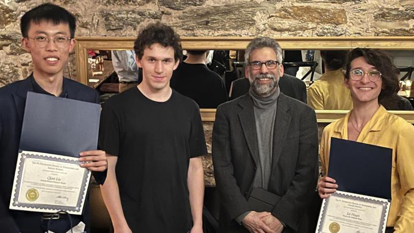 Four men stand in a row. Two are holding award certificates.