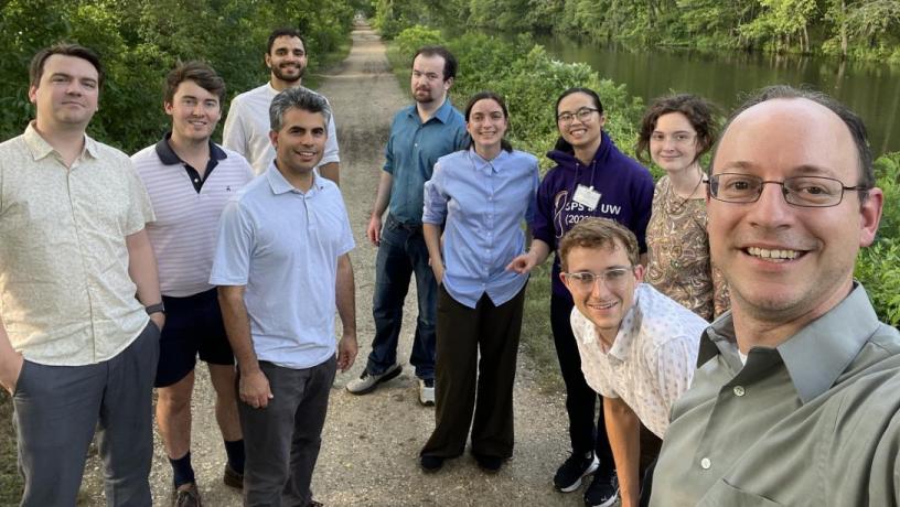 Students and faculty, both male and female, are standing outdoors in front of trees, a stream, and a walking path