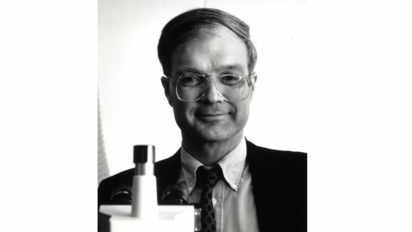 A middle aged white man is standing behind a microscope. He is wearing a suit coat and tie.