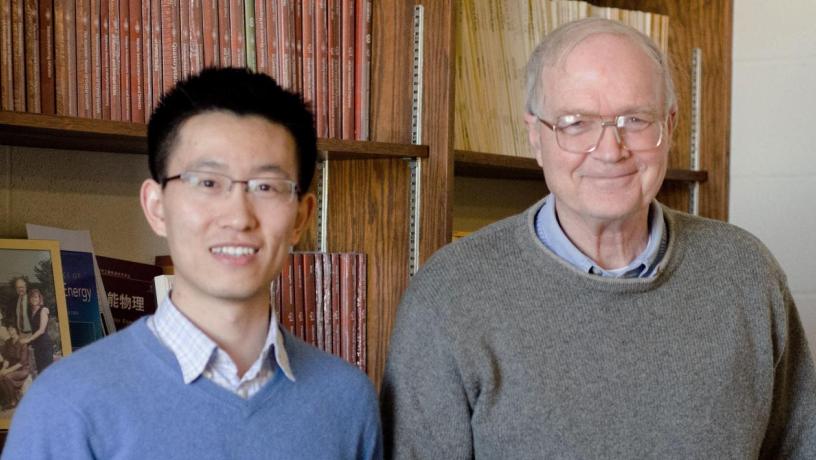Two men are standing in front of a bookshelf and smiling at the camera. One is a young Asian man with dark hair who is wearing a blue sweater. The other man is an elderly white person with white hair who is wearing a grey sweater.