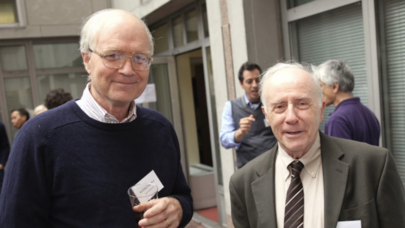 Two elderly white men are standing outdoors and are smiling at the camera. One man has white hair, glasses, and is wearing a dark blue sweater. The other man also has white hair and is wearing a grey jacket and striped tie.