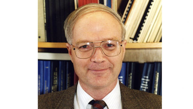 A middle aged white man is standing in front of a bookshelf. He is wearing a suit coat and tie.