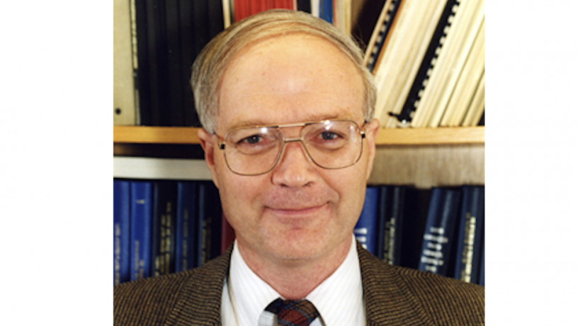 A middle aged white man is standing in front of a bookshelf. He is wearing a suit coat and tie. 