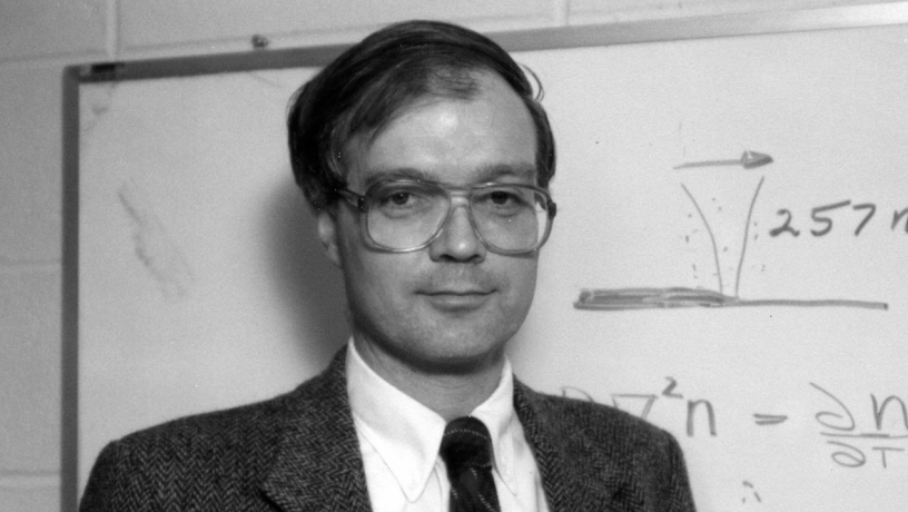 A middle aged white man standing in front of a black board. He is wearing a suit coat and tie.