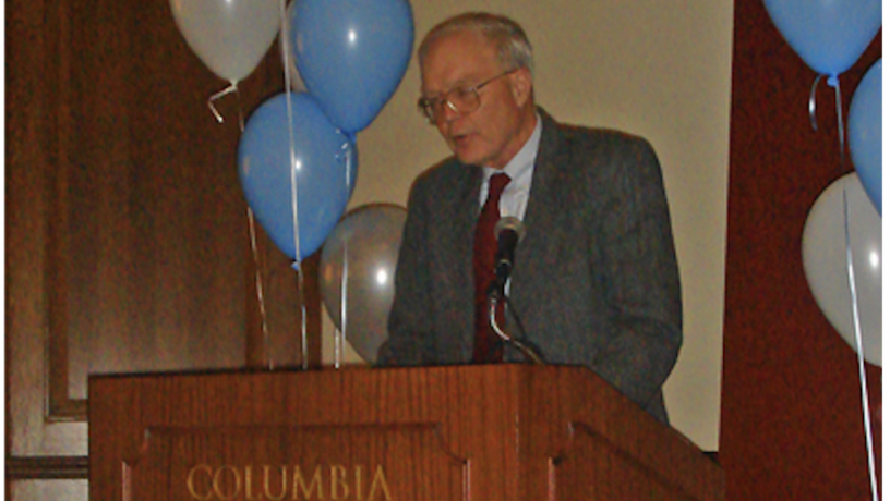 An elderly white man with white hair. He is wearing glasses, a grey suit coat, and a maroon tie. He is standing at a wood podium and is surrounded by blue and white balloons.