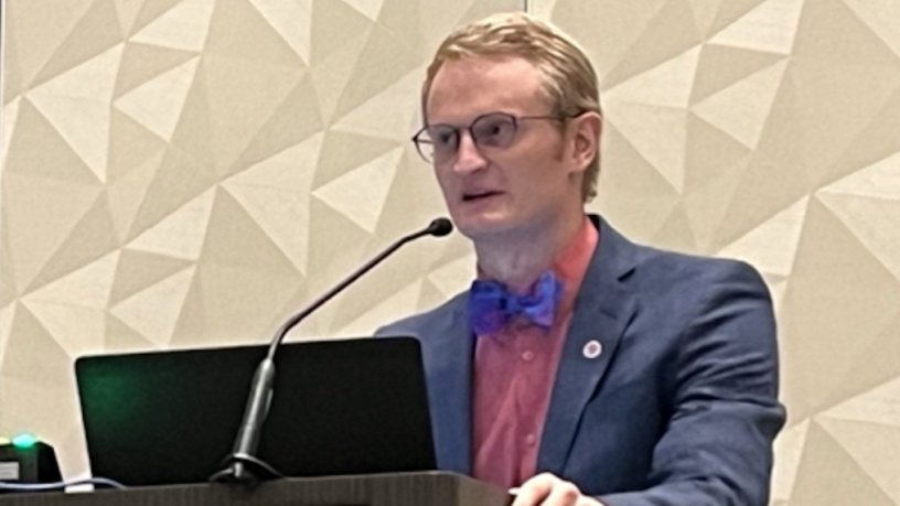 A young white male researcher stands at a podium. He has blond hair and glasses. He is wearing a blue jacket and bowtie.