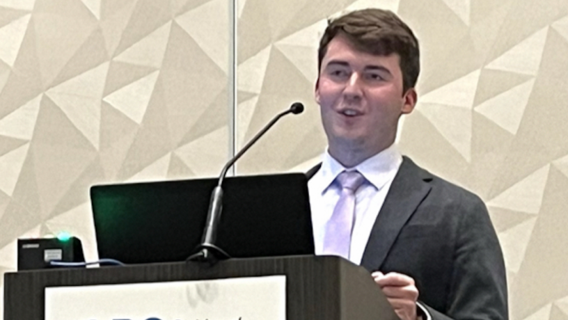 A young white male student stands at a podium. He has dark brown hair and is wearing a grey jacket and violet tie.