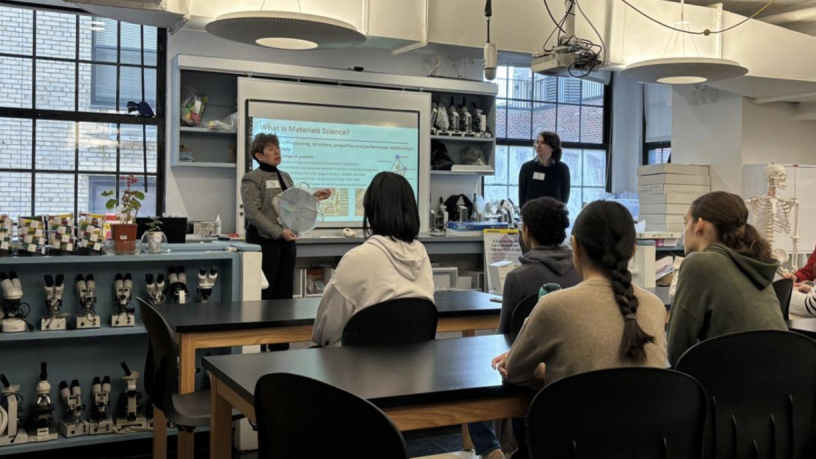 A woman stands in front of a large lab classroom full of four female students and one male student.
