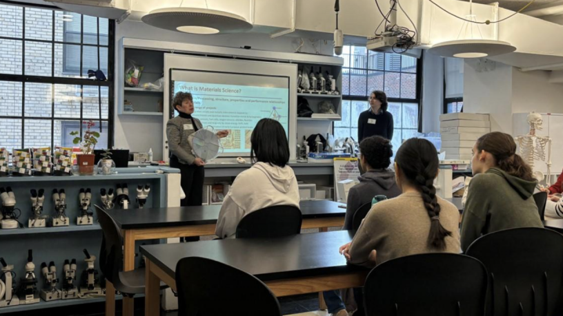 A woman stands in front of a large lab classroom full of four female students and one male student. 