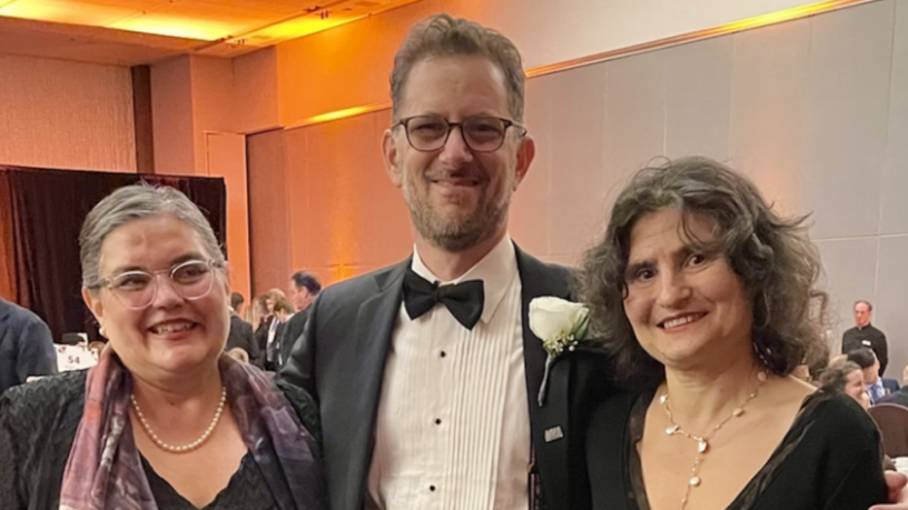 Three people stand in a ball room. A woman, a man, and another women are wearing formal wear.