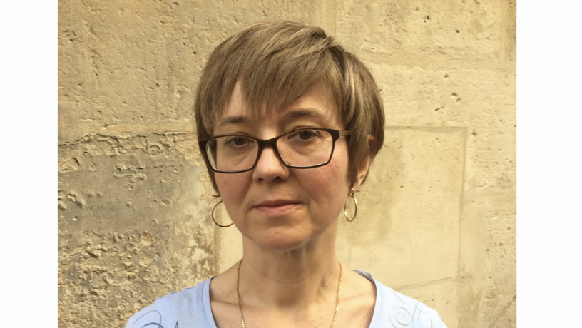 A photo of a white woman with light brown hair. She is wearing glasses and a light blue shift.