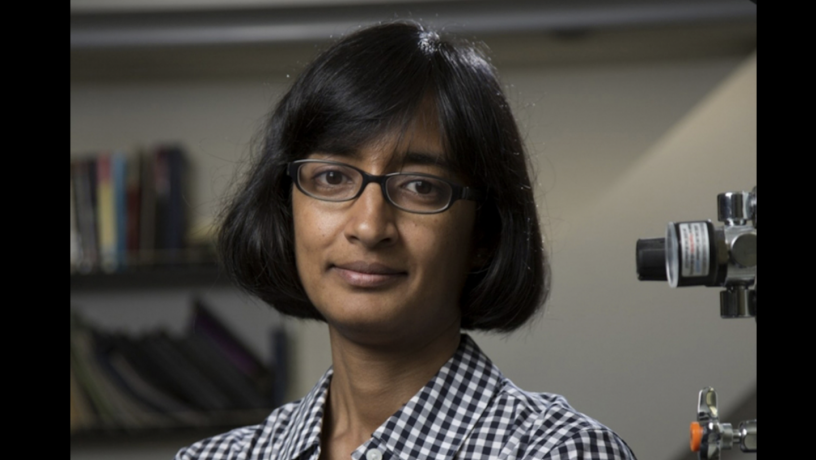 A photo of a woman standing in a laboratory. She has short black hair and is wearing glasses and a checkered shirt.