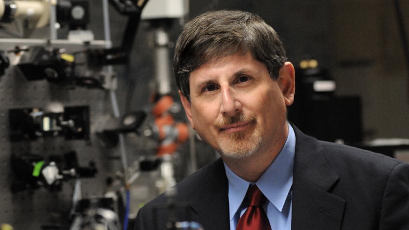 A photo of a white man with brown and gray hair. He is sitting in a lab and wearing a suit coat and tie.