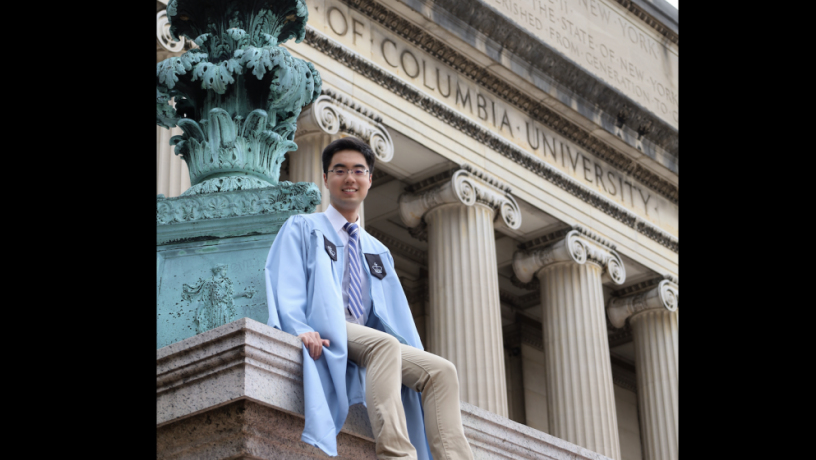 A photo of a young man with dark hair and glasses sitting outdoors wearing a light blue graduation gown