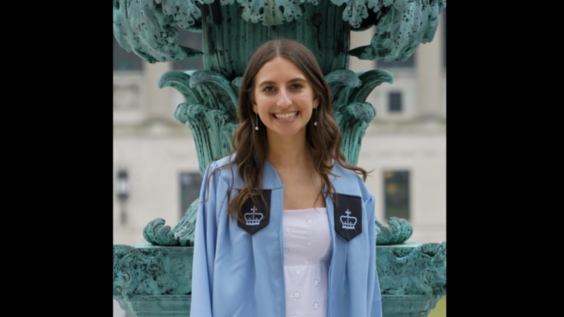 A photo of a young woman with long brown hair. She is standing outside and wearing a light blue graduation gown