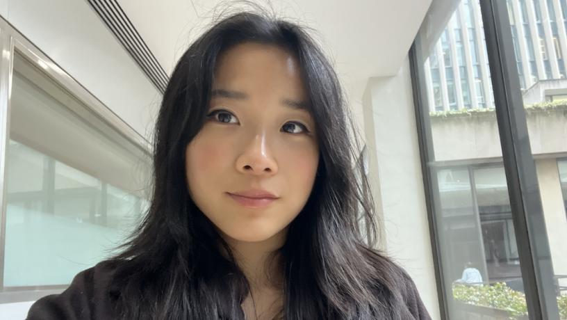 A photo of a young woman with black hair. She is wearing a black top and standing next to a window.