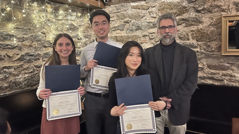 Three students and one faculty member stand in a restaurant. The students are holding up award certificates.