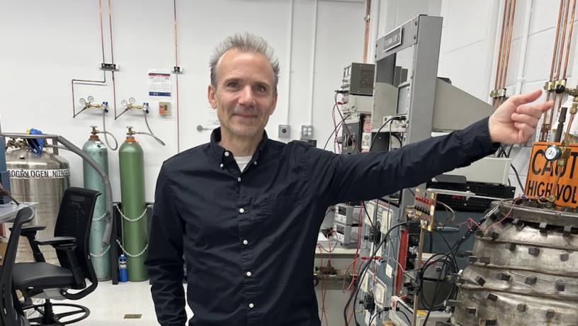 A photo of a man standing in a lab. He is wearing a black shirt and is pointing to machinery