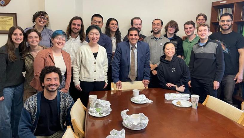A large group of college students and a middle aged man stand around a conference room table