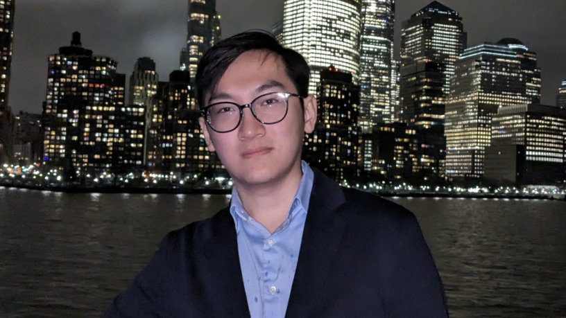 A young man stand outside infront of the NYC skyline. He is wearing a light blue shirt and navy blazer.