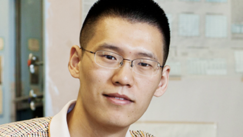 A photo a young Asian man with short black hair and glasses. He is sitting in a lab and looking at the camera.