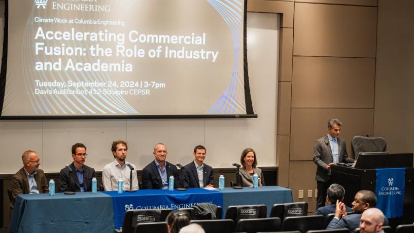 Six people sit at a panelist table at the front of a large auditorium. There is a large projection screen behind them. Another man stands at a podium and speaks into a microphone.