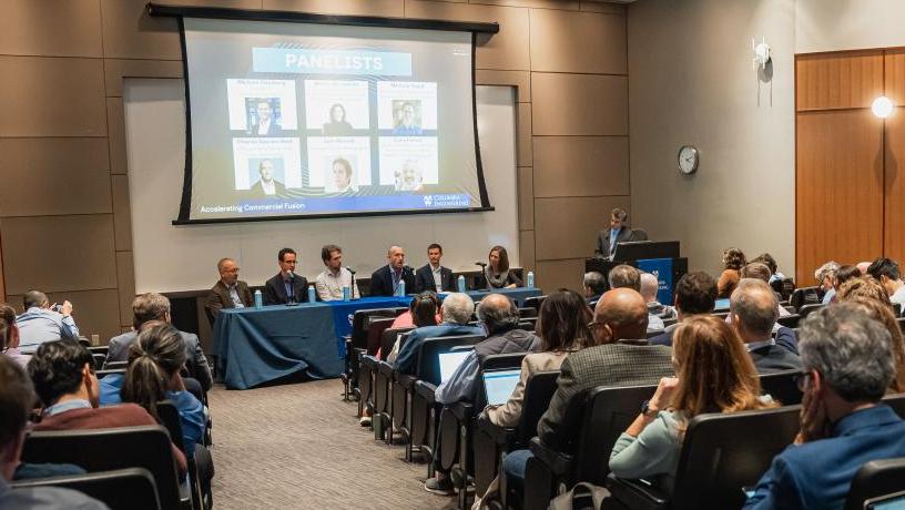 people in in an auditorium facing a table of panelists and a projection screen