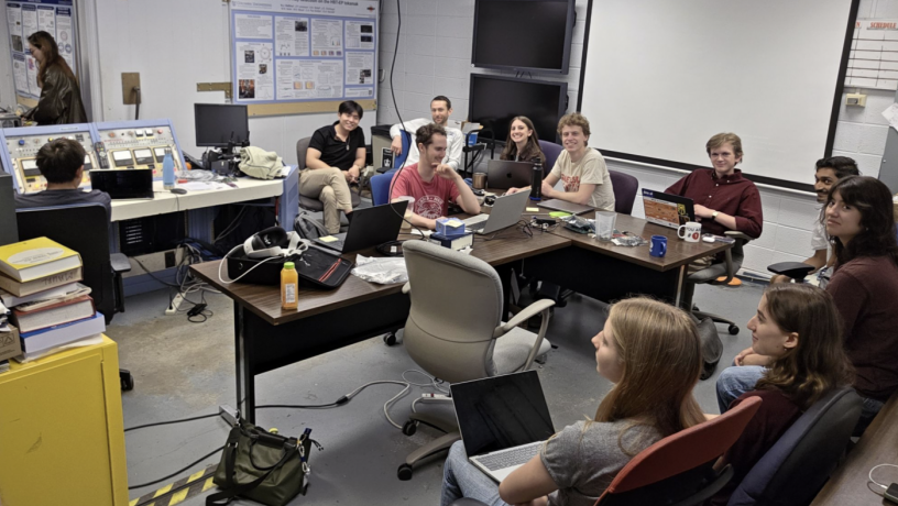 A group of students are meeting in a lab room with tables and computers