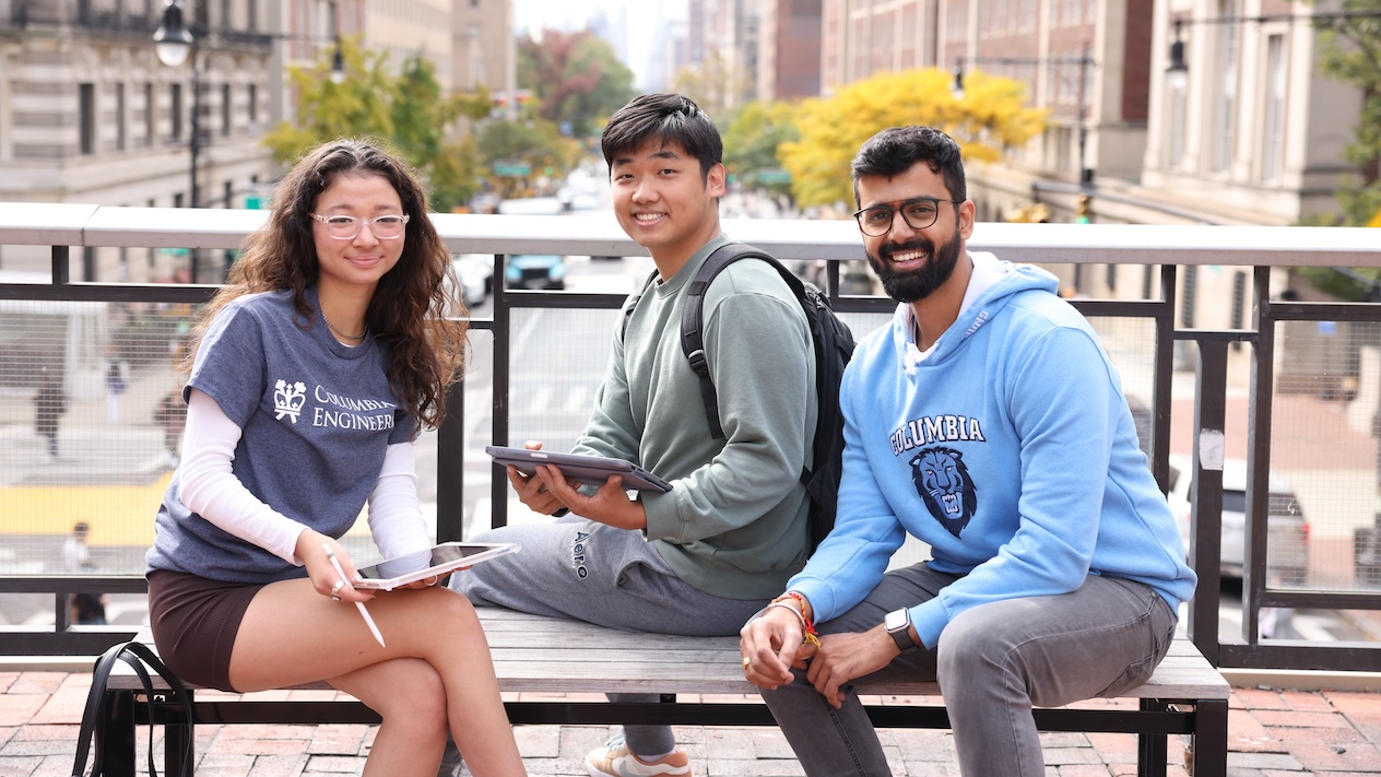 Three Columbia Engineering students smiling for a photo while sitting on a bench on Columbia ccampus. Two of the students are holding tablet computers