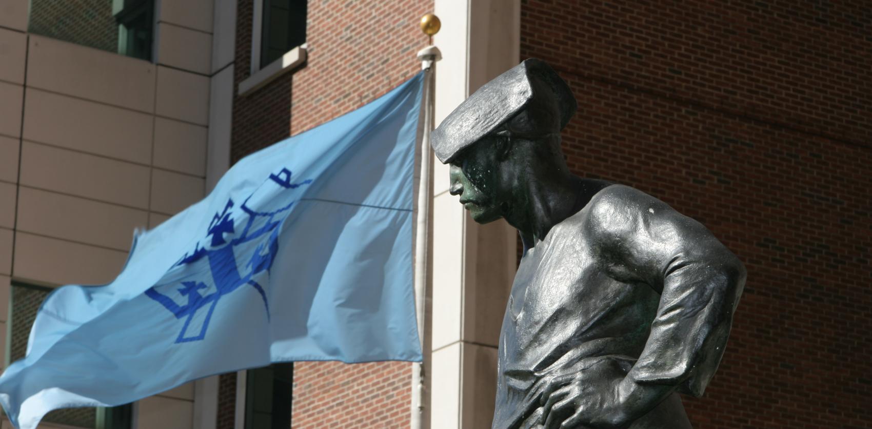 Bronze statue of a man wearing a cap, looking downward with hands on hips, in front of a brick building with a blue flag waving on a pole nearby.