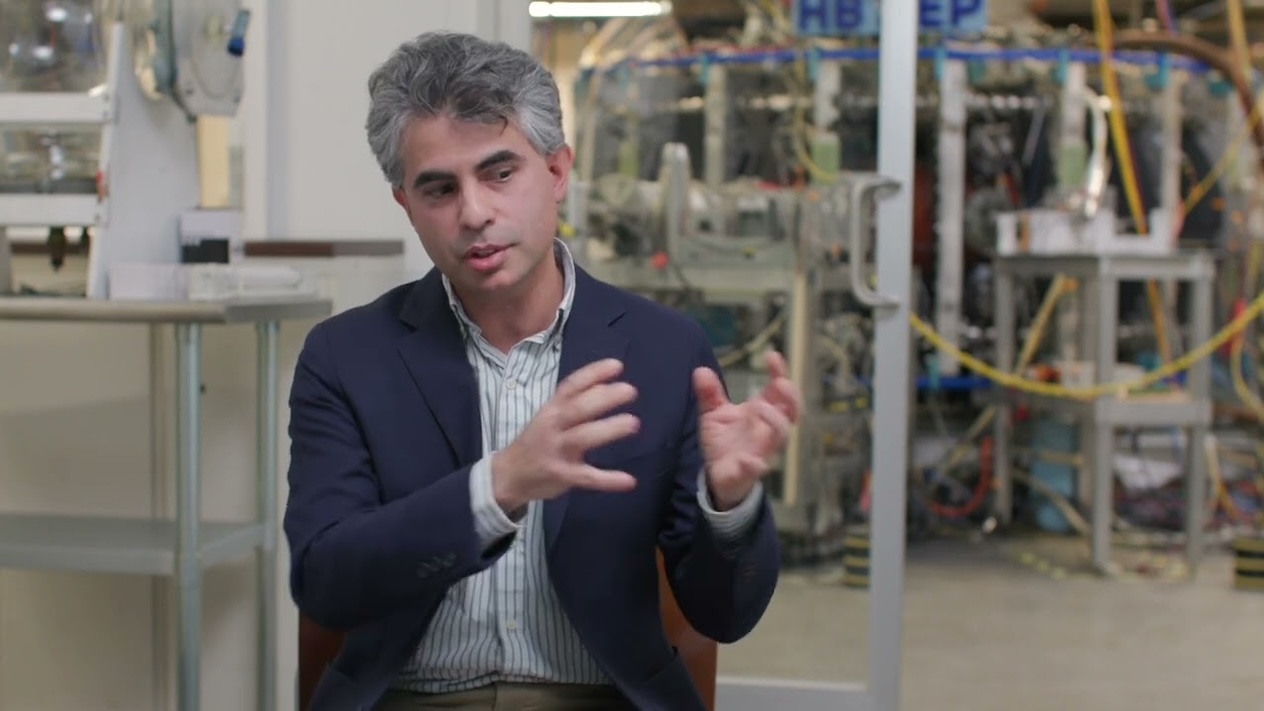 Carlos Paz-Soldan gesturing. Behind him, blurred lab equipment and cables are visible through glass panels.