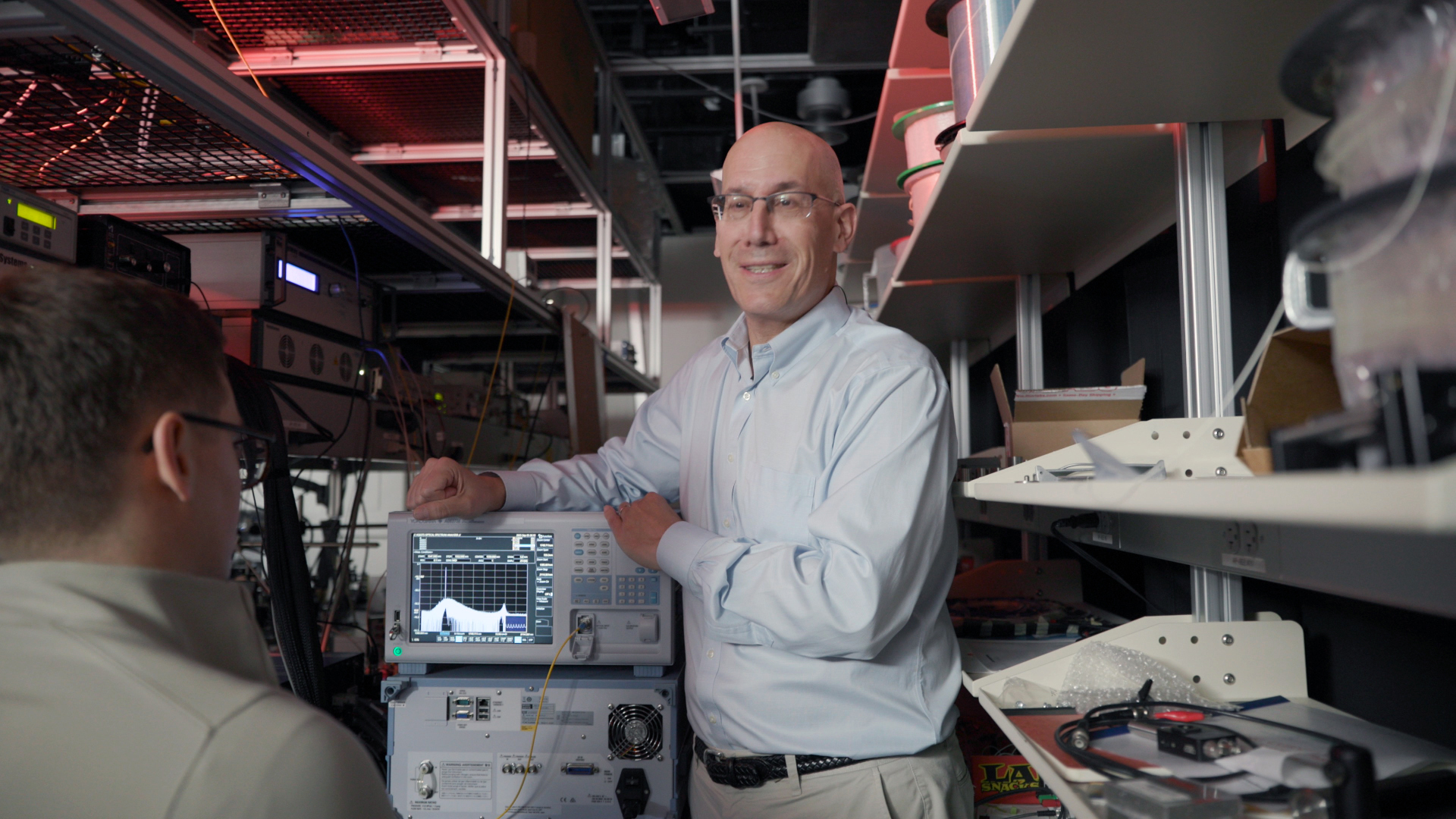 Alexander Gaeta smiles while standing beside lab equipment with a spectrum display on a monitor, speaking to another person seated in the foreground in a cluttered electronics lab.