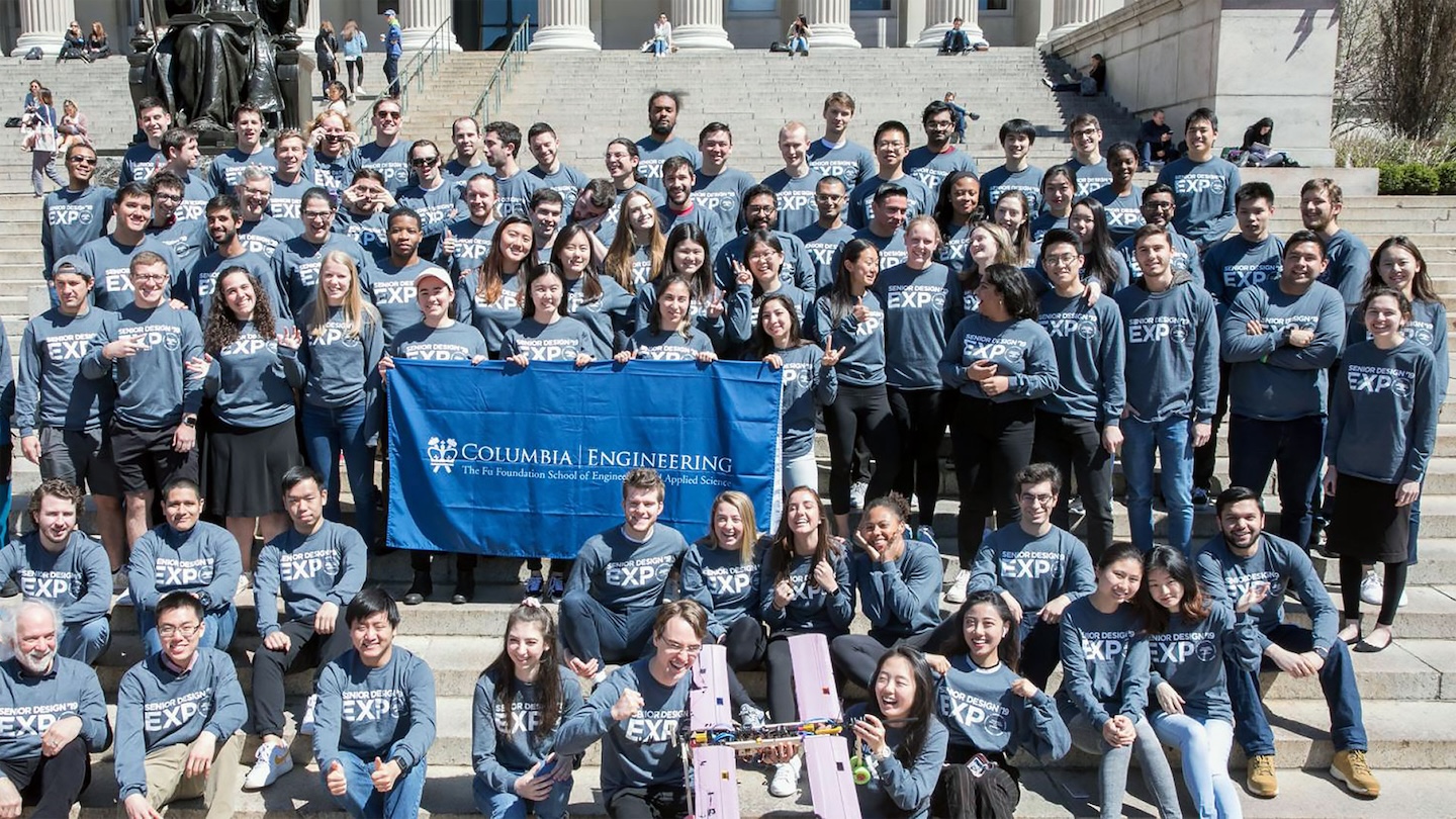 a large group of students wearing shirts that say 2019 senior design expo sit outside on the steps in front of Columbia's Low Library and a statue of alma mater