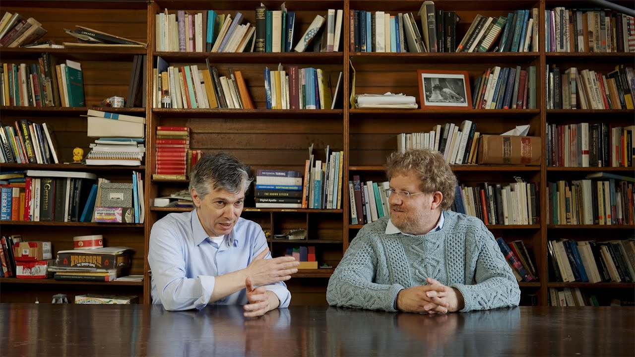 Two men sit at a dark table in conversation in front of a large wooden bookshelf filled with books and academic materials. The man on the left wears a light blue shirt, and the man on the right wears a gray knit sweater.
