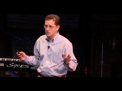 Man in a light blue button-up shirt presents on stage with animated hand gestures while speaking. A keyboard is visible in the background.