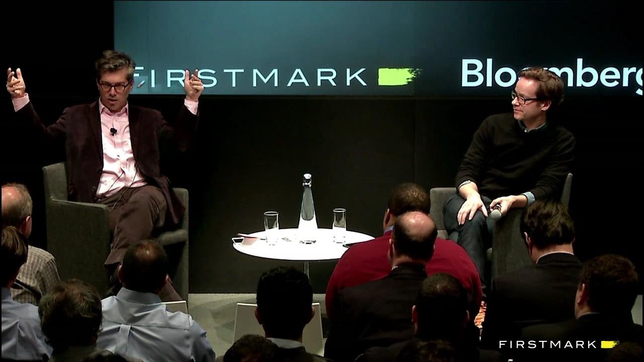Two men sit in chairs engaged in discussion at a round white table on stage during a FirstMark Bloomberg event, with audience members seated in the foreground.