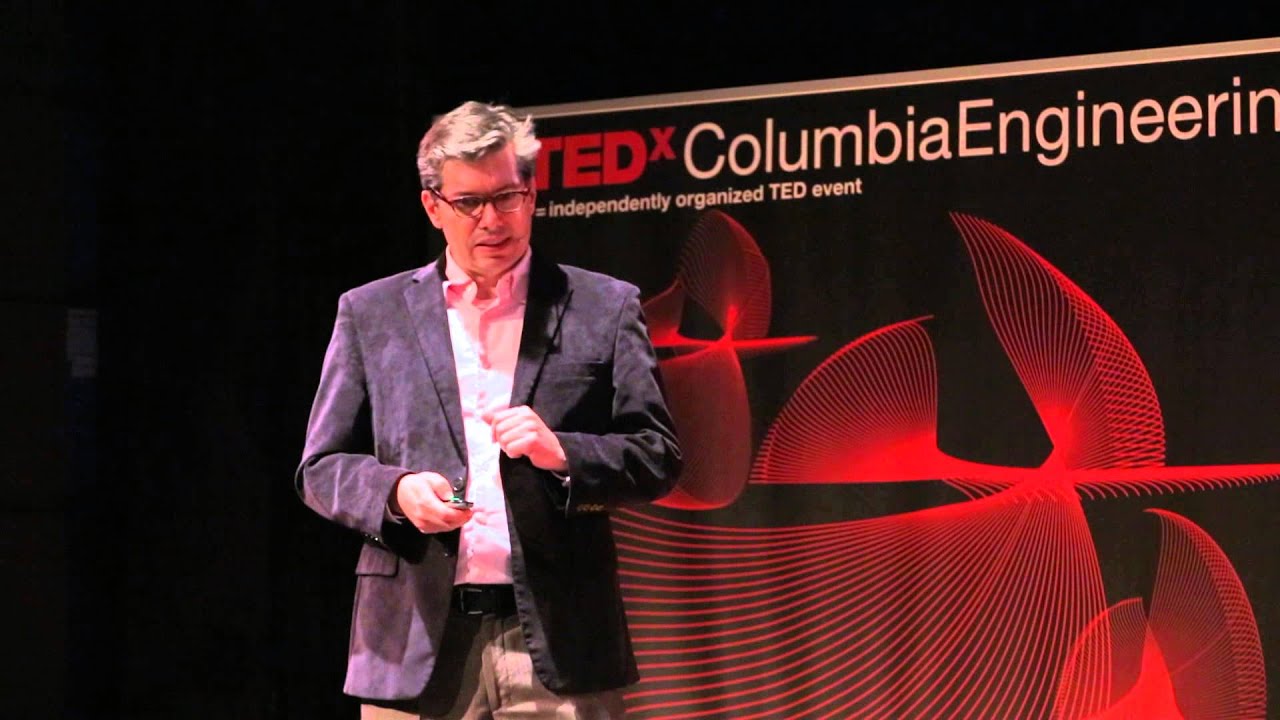Speaker in a gray blazer and white shirt holds a microphone while presenting on stage at a TEDx Columbia Engineering event, with the event branding visible on the backdrop behind him.