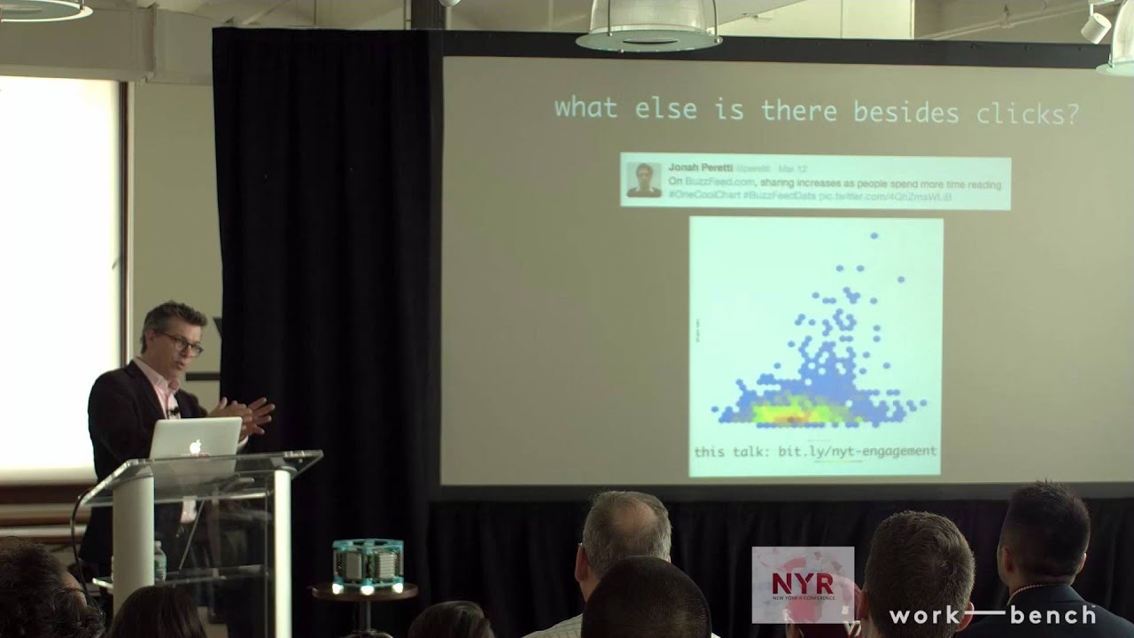 Man in glasses and dark blazer speaks at a podium with a laptop during a presentation. A projection screen behind him displays text reading "what else is there besides clicks?" and shows a scatter plot data visualization. The audience is partially visible in the foreground.