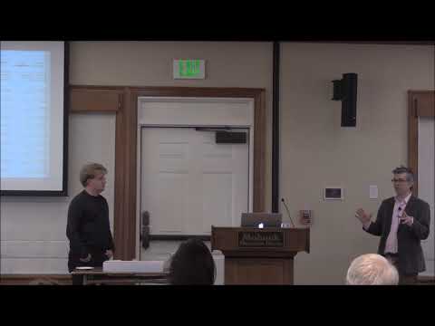 Academic seminar scene with a woman standing on the left and a man in business attire gesturing while speaking on the right in front of a whiteboard in an institutional setting.
